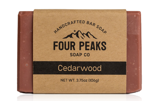 A bar of handcrafted soap from Four Peaks Soap Co. labeled "Cedarwood." The soap is wrapped in a brown paper band with a mountain logo and text indicating it weighs 3.75 ounces (106 grams).