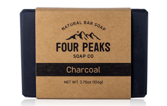 A bar of handcrafted soap from Four Peaks Soap Co. labeled "Charcoal." The soap is wrapped in a brown paper band with a mountain logo and text indicating it weighs 3.75 ounces (106 grams).
