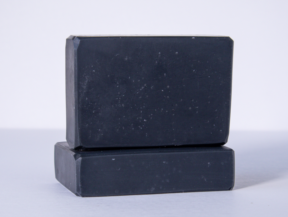 Two black bars of soap are stacked on a clean white surface with one bar lying flat and the other placed upright on top. The soaps have a smooth texture and rectangular shape with slightly rounded edges.