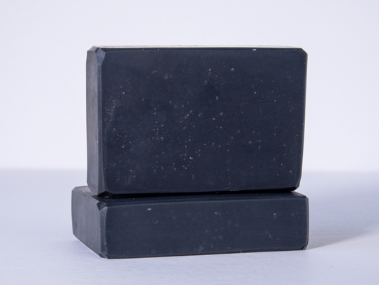 Two black bars of soap are stacked on a clean white surface with one bar lying flat and the other placed upright on top. The soaps have a smooth texture and rectangular shape with slightly rounded edges.