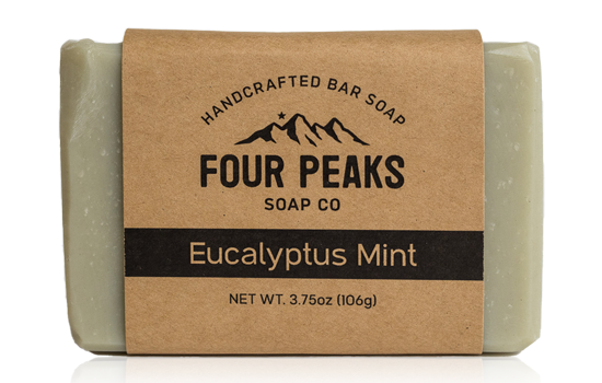 A bar of handcrafted soap from Four Peaks Soap Co. labeled "Eucalyptus Mint." The soap is wrapped in a brown paper band with a mountain logo and text indicating it weighs 3.75 ounces (106 grams).