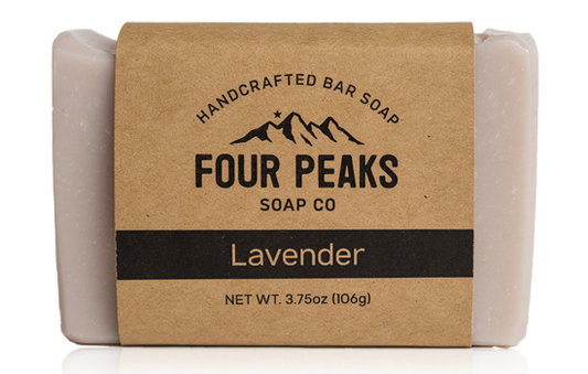 A bar of handcrafted soap from Four Peaks Soap Co. labeled "Lavender." The soap is wrapped in a brown paper band with a mountain logo and text indicating it weighs 3.75 ounces (106 grams).