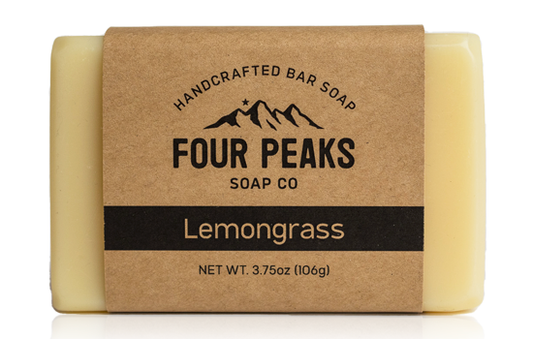 A bar of handcrafted soap from Four Peaks Soap Co. labeled "Lemongrass." The soap is wrapped in a brown paper band with a mountain logo and text indicating it weighs 3.75 ounces (106 grams).