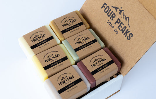 A cardboard box labeled "Four Peaks Soap Co." containing six different bars of soap, each individually wrapped in brown paper. The soaps are labeled as Bare Naked, Oatmeal Honey, Eucalyptus Mint, Lemongrass, Lavender, and Cedarwood
