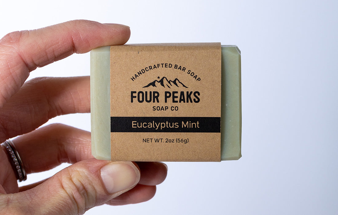 A hand holding a bar of handcrafted soap wrapped in brown paper. The label reads: "Four Peaks Soap Co" with a mountain logo above it. Beneath, it says "Eucalyptus Mint" and "NET WT. 2oz (56g)."