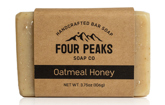 A bar of handcrafted soap from Four Peaks Soap Co. labeled "Oatmeal Honey." The soap is wrapped in a brown paper band with a mountain logo and text indicating it weighs 3.75 ounces (106 grams).