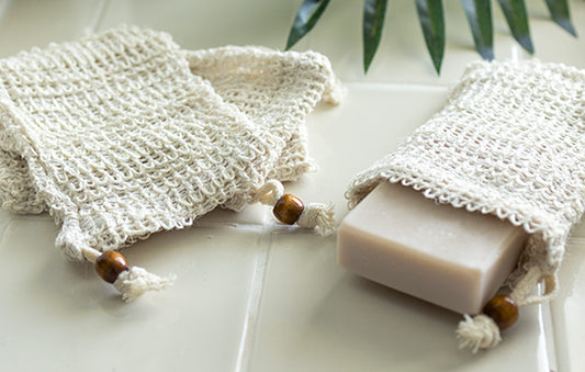 Two natural soap saver bags, each holding a bar of soap, lie on a white tiled surface. The bags have a woven texture with drawstrings adorned with wooden beads.