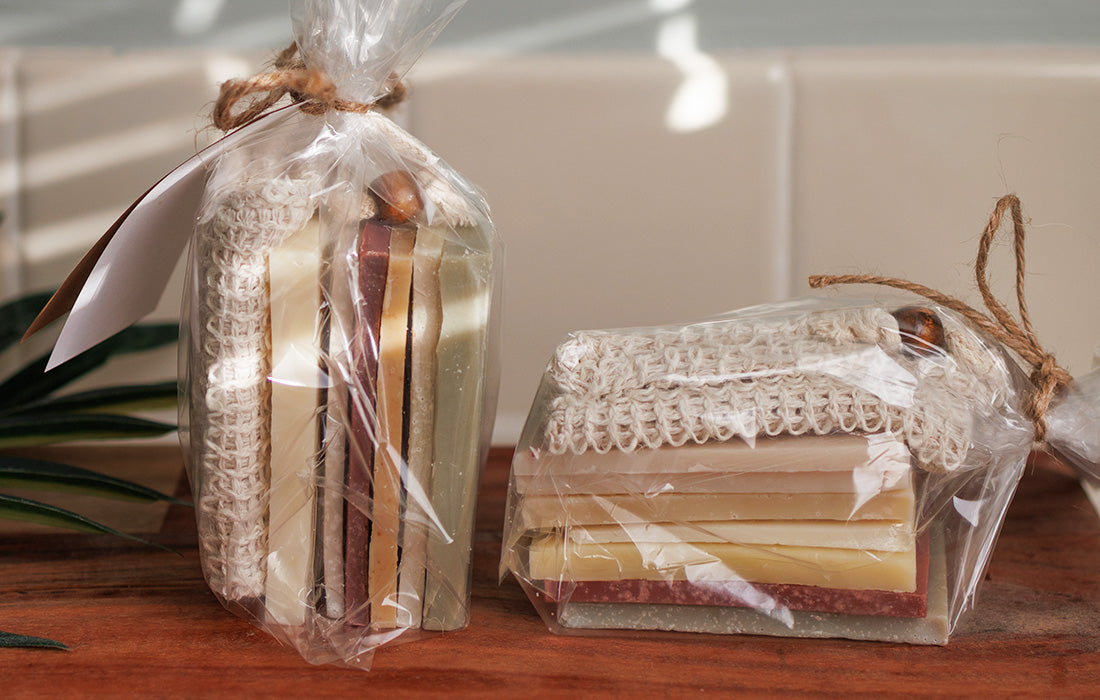 Two gift sets of handmade soap bars wrapped in clear plastic with twine bows sit on a wooden surface. Each set appears to include multiple soap bars in various colors and a scrubby cloth.