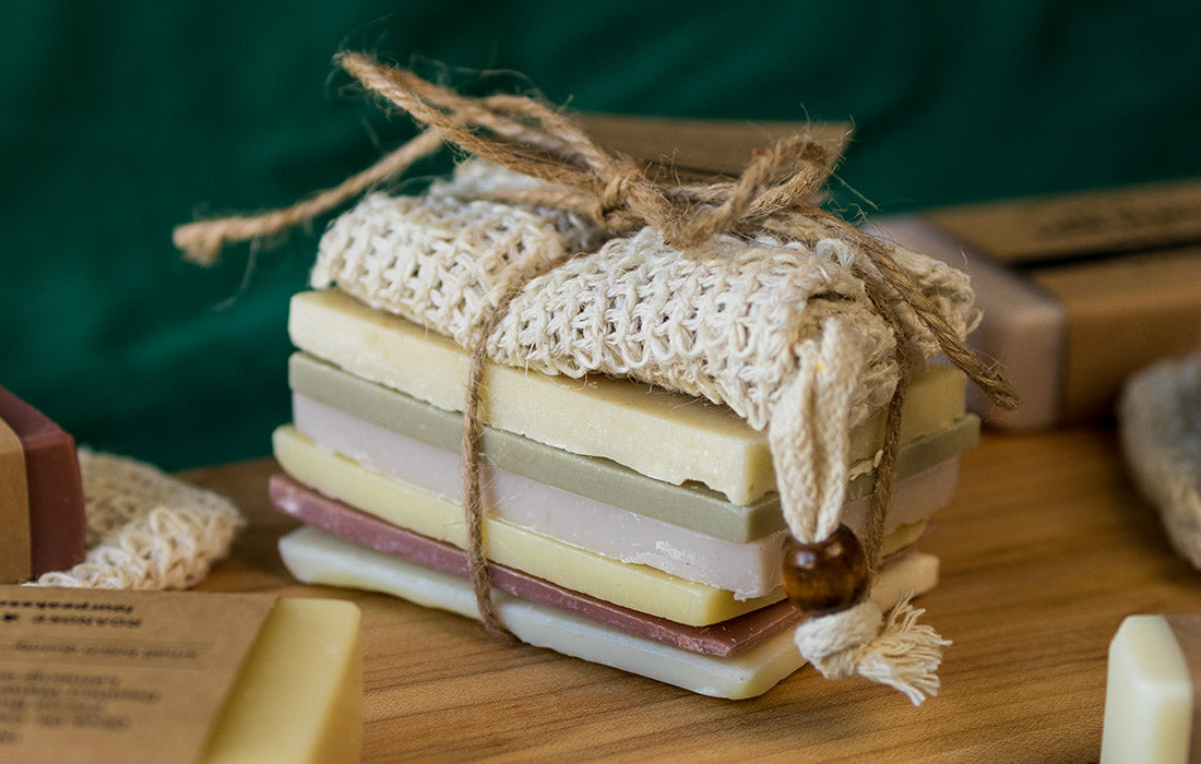 A stack of colorful soap bars are tied together with a piece of twine. They are arranged neatly on a wooden surface, accompanied by a soap saver bag and a small brown bead for decoration. The background is slightly blurred but shows a green fabric.