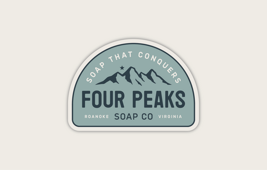 A sticker logo for Four Peaks Soap Co. features an illustration of four mountain peaks with a star above them. Text around the top reads "Soap That Conquers." Below the peaks, it says "Four Peaks Soap Co" with "Roanoke Virginia" underneath. The logo has a blue and gray color scheme.