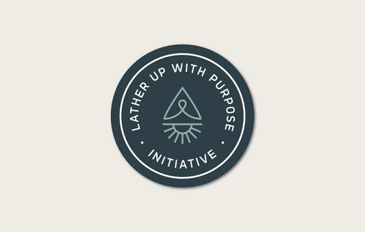 A sticker of the "Lather Up With Purpose" Initiative. The logo features a circular shape with text that reads "Lather Up With Purpose Initiative". Inside the circle there is an icon of sun, cancer ribbon, and compass arrow that when combined represents a water drop. The logo has a blue and teal color scheme.